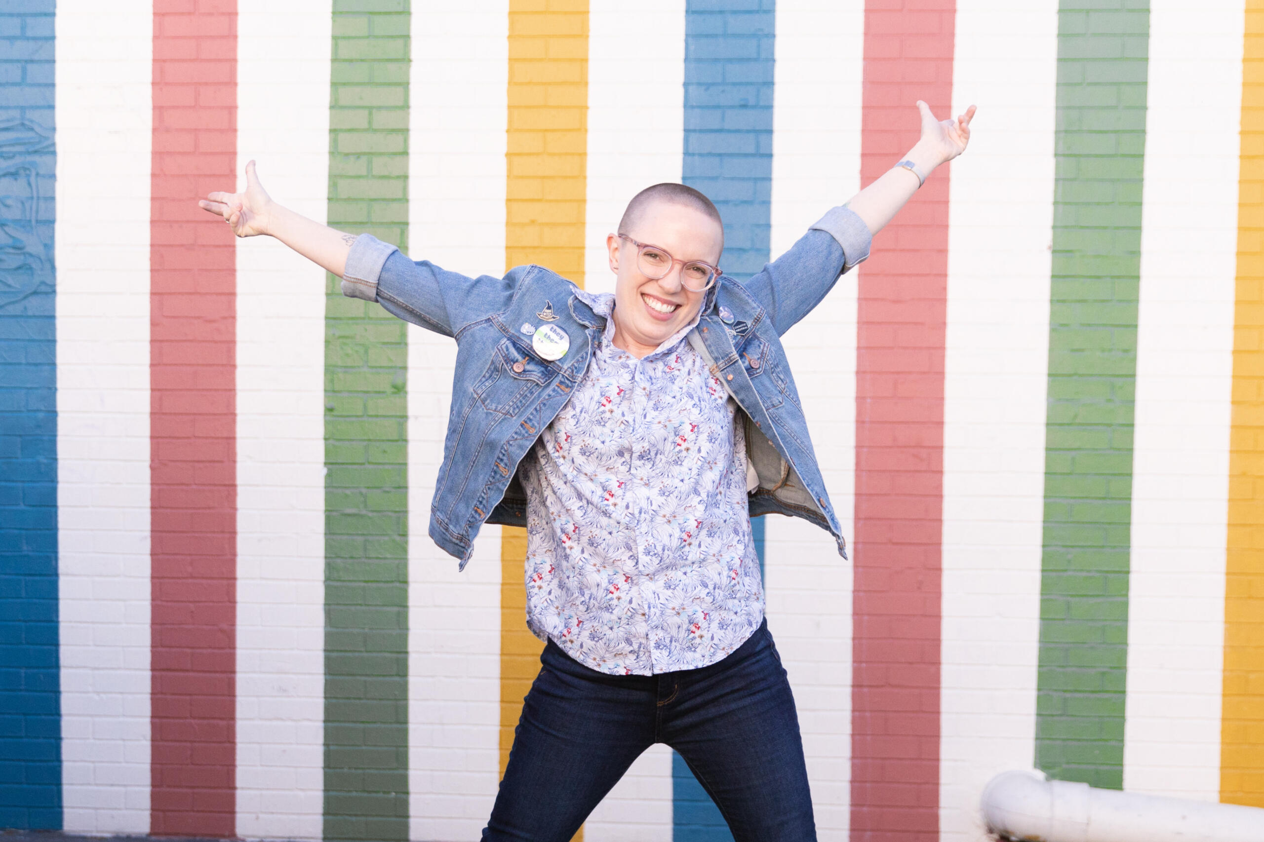 Anna wearing glasses, a button down shirt, and a jean jacket. They are standing in front of a rainbow striped wall.